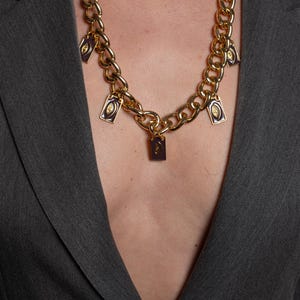 May include: A gold-toned chunky chain necklace featuring five rectangular purple enamel charms. Each charm displays a centered gold eye motif. The necklace is worn over a dark gray blazer, with the chain links forming a prominent collar design.