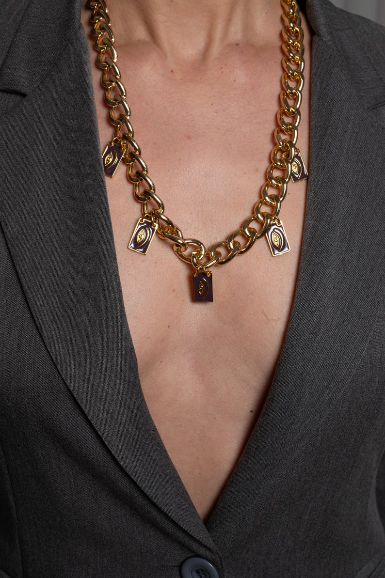 May include: A gold-toned chunky chain necklace featuring five rectangular purple enamel charms. Each charm displays a centered gold eye motif. The necklace is worn over a dark gray blazer, with the chain links forming a prominent collar design.