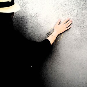 Puede incluir: Una persona con un sombrero de paja y una camisa negra con pulseras doradas en la mu&ntilde;eca, de pie frente a una pared blanca texturizada.