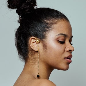 May include: A woman with dark hair pulled back in a bun wears a gold ear cuff with a dangling chain and a black bead.