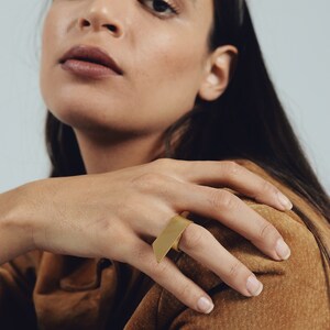 May include: A gold-toned ring featuring a flat, rectangular metal plate that extends horizontally across the finger. The surface of the rectangular plate is smooth and matte, creating a clean, geometric look that sits prominently on the hand.