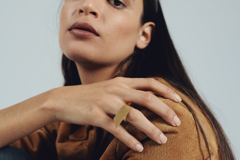 May include: A gold-toned ring featuring a flat, rectangular metal plate that extends horizontally across the finger. The surface of the rectangular plate is smooth and matte, creating a clean, geometric look that sits prominently on the hand.