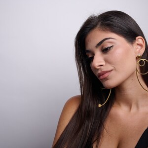 May include: A woman with long dark hair wearing a black top and large gold hoop earrings with a dangling circle design.