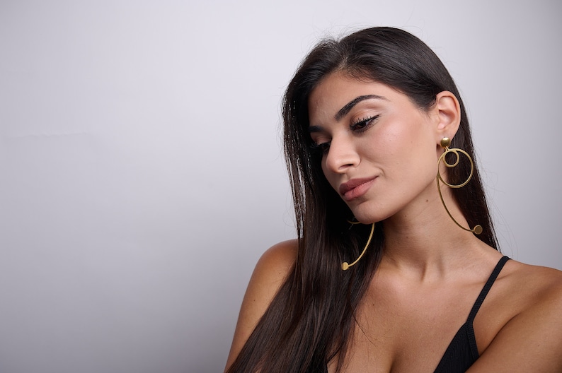 May include: A woman with long dark hair wearing a black top and large gold hoop earrings with a dangling circle design.