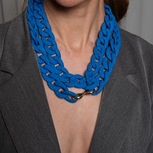 May include: A close-up of a woman wearing a blue chunky chain necklace. The necklace is made of thick, linked plastic and has a gold clasp.
