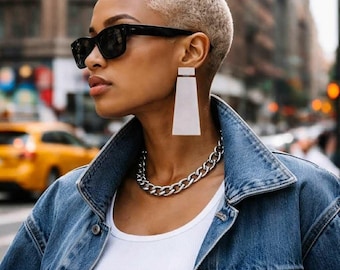 May include: A person with short blonde hair wearing black rectangular sunglasses, large white geometric drop earrings, a thick silver chain link necklace, a white tank top, and a blue denim jacket. The style is captured in an urban setting.