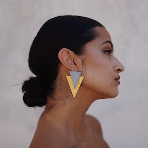 May include: A pair of geometric earrings featuring two overlapping triangles. The top triangle is a matte grey, while the larger bottom triangle is a polished gold tone. These earrings hang vertically from the earlobe, creating a layered effect.