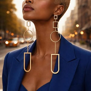 May include: Gold-toned geometric dangle earrings featuring squares, circles, and U-shaped pendants. The earrings are suspended from delicate chains. The model is wearing a blue blazer, with a blurred city street in the background.