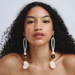 May include: A woman with long curly dark hair wearing large statement earrings with white pearls, brown and gold beads, and a white shell pendant.
