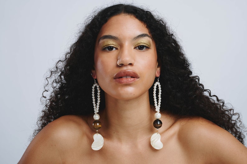 May include: A woman with long curly dark hair wearing large statement earrings with white pearls, brown and gold beads, and a white shell pendant.