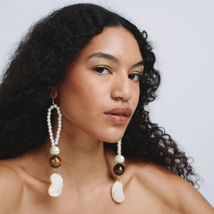 May include: A person with long curly dark hair wearing a pair of long pearl and shell earrings. The earrings have a gold accent and brown tiger eye beads.