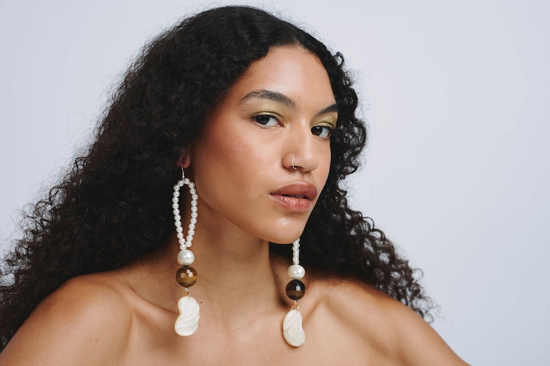 May include: A person with long curly dark hair wearing a pair of long pearl and shell earrings. The earrings have a gold accent and brown tiger eye beads.