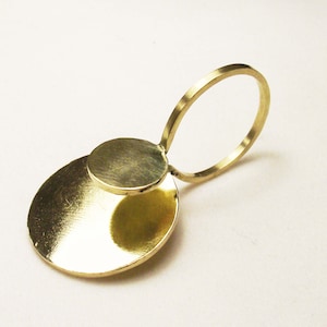 May include: A gold-toned ring featuring a thin, circular band attached to a larger, flat, circular disc element. The metal surface has a polished, reflective finish, and the design consists of two distinct circular shapes connected together.