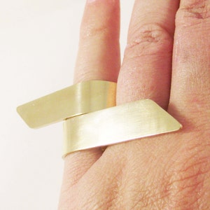 May include: A gold-toned ring featuring two overlapping, flat, rectangular metal bands that wrap around the finger. The surface of the metal has a brushed, matte finish with angled, pointed ends on each of the two parallel, horizontal segments.