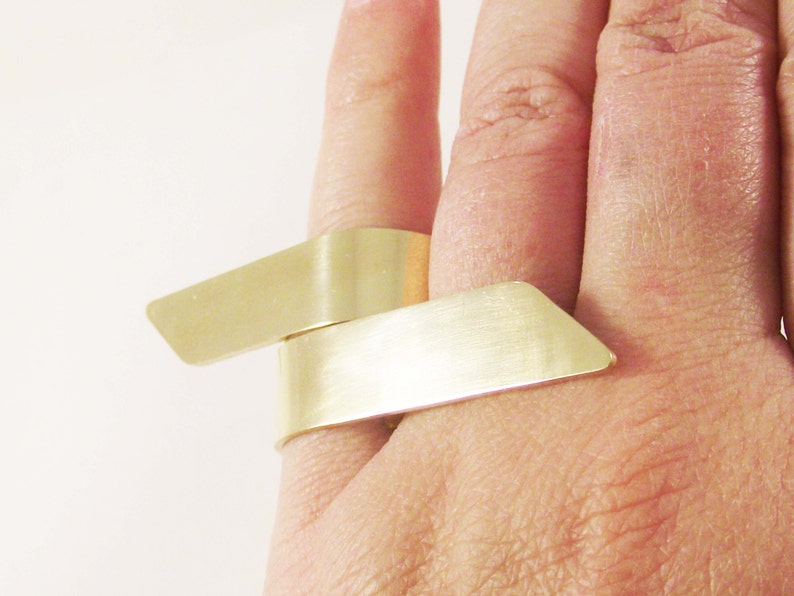 May include: A gold-toned ring featuring two overlapping, flat, rectangular metal bands that wrap around the finger. The surface of the metal has a brushed, matte finish with angled, pointed ends on each of the two parallel, horizontal segments.