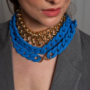 May include: A layered necklace with a thick blue plastic chain and a gold chain. The necklace is worn by a person with a gray blazer.