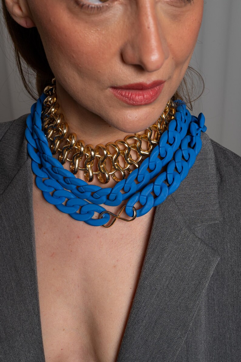 May include: A layered necklace with a thick blue plastic chain and a gold chain. The necklace is worn by a person with a gray blazer.