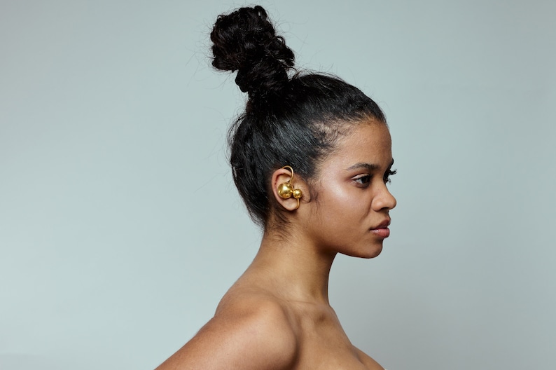May include: A woman with dark hair styled in a high bun wears a gold ear cuff with two round, gold spheres.