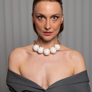 May include: A statement necklace featuring large, smooth, spherical white beads separated by small gold-toned metal spacers, connected to a gold-toned chain. The necklace is worn with a dark gray blazer that has an off-the-shoulder neckline.
