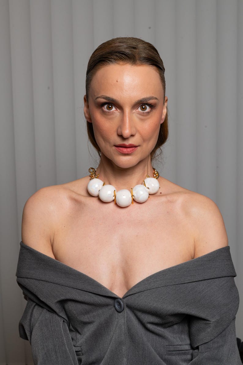 May include: A statement necklace featuring large, smooth, spherical white beads separated by small gold-toned metal spacers, connected to a gold-toned chain. The necklace is worn with a dark gray blazer that has an off-the-shoulder neckline.