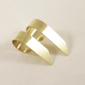 May include: Two gold-colored metal rings with a smooth, polished finish. Each ring features a wide, curved band that transitions into a flat, pointed extension, creating a distinct angular shape that rests on a plain, light-colored surface.