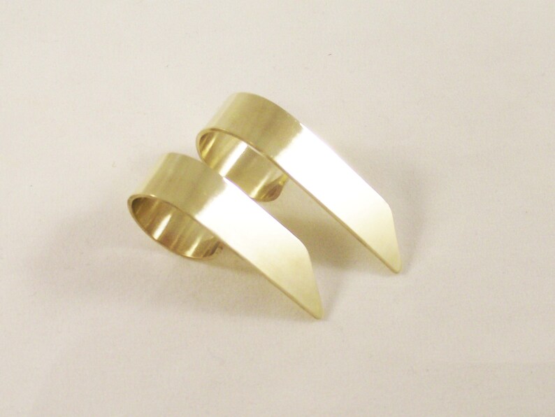 May include: Two gold-colored metal rings with a smooth, polished finish. Each ring features a wide, curved band that transitions into a flat, pointed extension, creating a distinct angular shape that rests on a plain, light-colored surface.