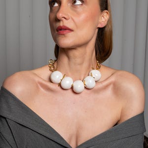 May include: A statement necklace featuring five large, smooth, white spherical beads separated by thin gold-toned metal bars. The beads are connected to a thick gold-toned chain link structure that rests around the neck, creating a bold contrast.