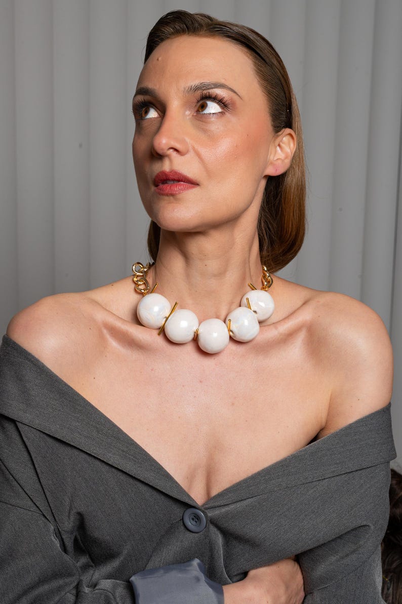 May include: A statement necklace featuring five large, smooth, white spherical beads separated by thin gold-toned metal bars. The beads are connected to a thick gold-toned chain link structure that rests around the neck, creating a bold contrast.