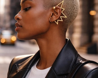Può includere: Un ear cuff color oro caratterizzato da una cornice geometrica a zigzag che si estende lungo il bordo esterno dell'orecchio, impreziosito da una piccola perla sferica liscia sulla parte superiore. Il pezzo è realizzato in filo metallico sottile.