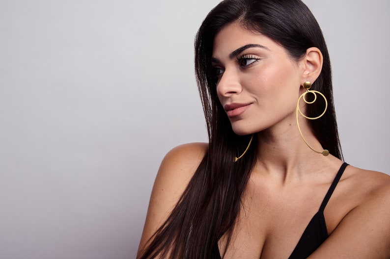 Model wearing long geometric gold statement earrings with circular design, modern sculptural dangle earrings in gold plated brass, fashion forward minimalist jewelry.