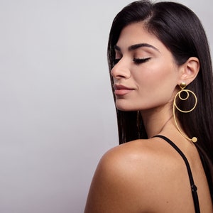 May include: A woman with long dark hair wearing a black vest top and gold hoop earrings with a dangling circle design.