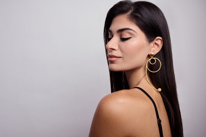 May include: A woman with long dark hair wearing a black vest top and gold hoop earrings with a dangling circle design.