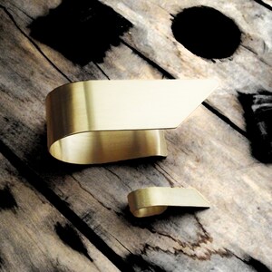 May include: Two gold-toned metal cuffs with a smooth, brushed finish. The larger cuff features a wide, curved band that tapers to an angled edge, while the smaller cuff mirrors this design in a compact size, both showcasing a clean, metallic surface.