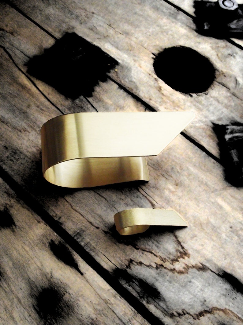 May include: Two gold-toned metal cuffs with a smooth, brushed finish. The larger cuff features a wide, curved band that tapers to an angled edge, while the smaller cuff mirrors this design in a compact size, both showcasing a clean, metallic surface.