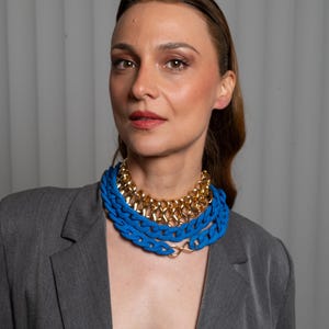 May include: A woman wearing a gray blazer with a layered necklace featuring a gold chain and a thick blue chain.
