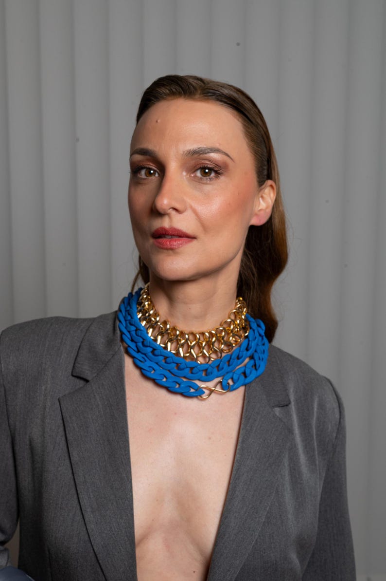 May include: A woman wearing a gray blazer with a layered necklace featuring a gold chain and a thick blue chain.