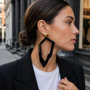 May include: A large, black, geometric-shaped earring with a gold-toned ball stud. The earring features an open, irregular, angular frame design. The piece is worn on an earlobe, paired with a white t-shirt and a black blazer, with a gold ring on a finger.