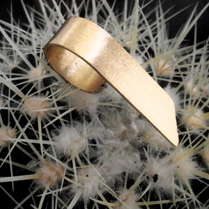 May include: A gold-toned metal ring with a wide, flat, and slightly angled band. The ring features a smooth, polished surface that reflects light, creating a metallic sheen. The band is open-ended, forming a continuous loop with a pointed end.