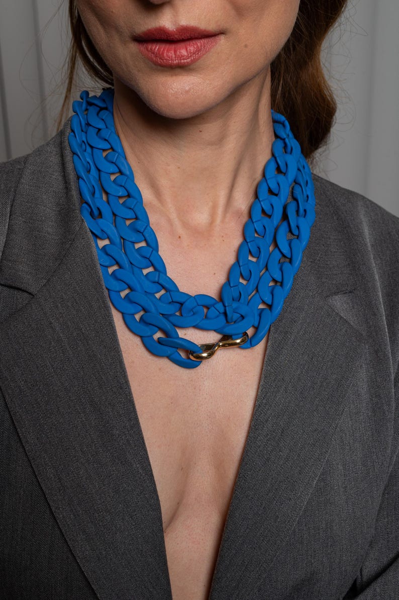 May include: A woman wearing a blue blazer with a thick, blue plastic chain necklace. The necklace is made of large, linked chain pieces.