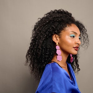 May include: A woman with long, curly black hair wearing a blue shirt and pink geometric earrings. She is looking to the right of the frame.