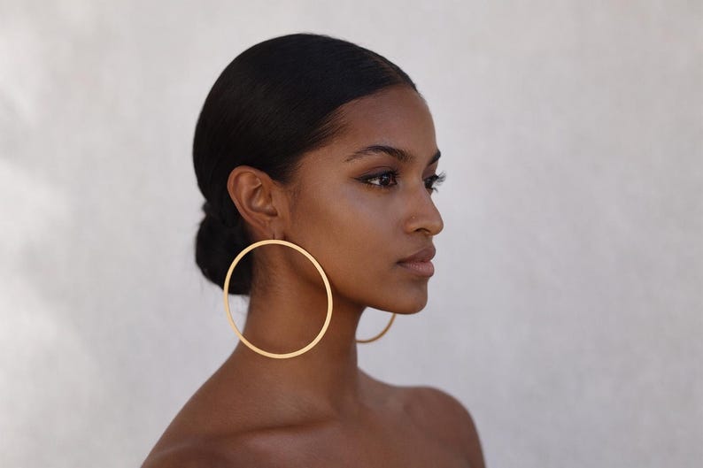 A side profile of a person with dark hair pulled back into a sleek low bun, showcasing a pair of oversized, thin gold hoop earrings. The earrings have a flat, brushed metallic finish and a large diameter that reaches down toward the shoulder. The person has a minimal makeup look with winged eyeliner, posed against a soft, neutral-colored background.