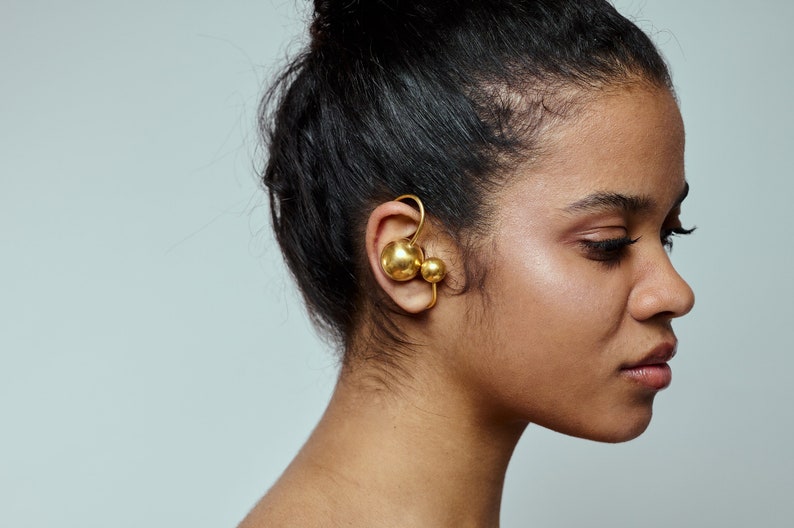 May include: A gold ear cuff with two round spheres. The cuff is worn on the left ear of a person with dark hair.