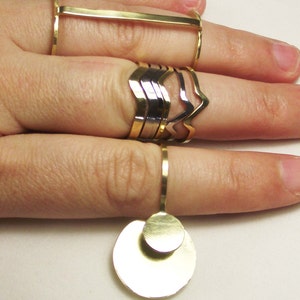 May include: A set of gold and silver rings, including a ring with a large, round, double-layered pendant. The rings are stacked on a finger.