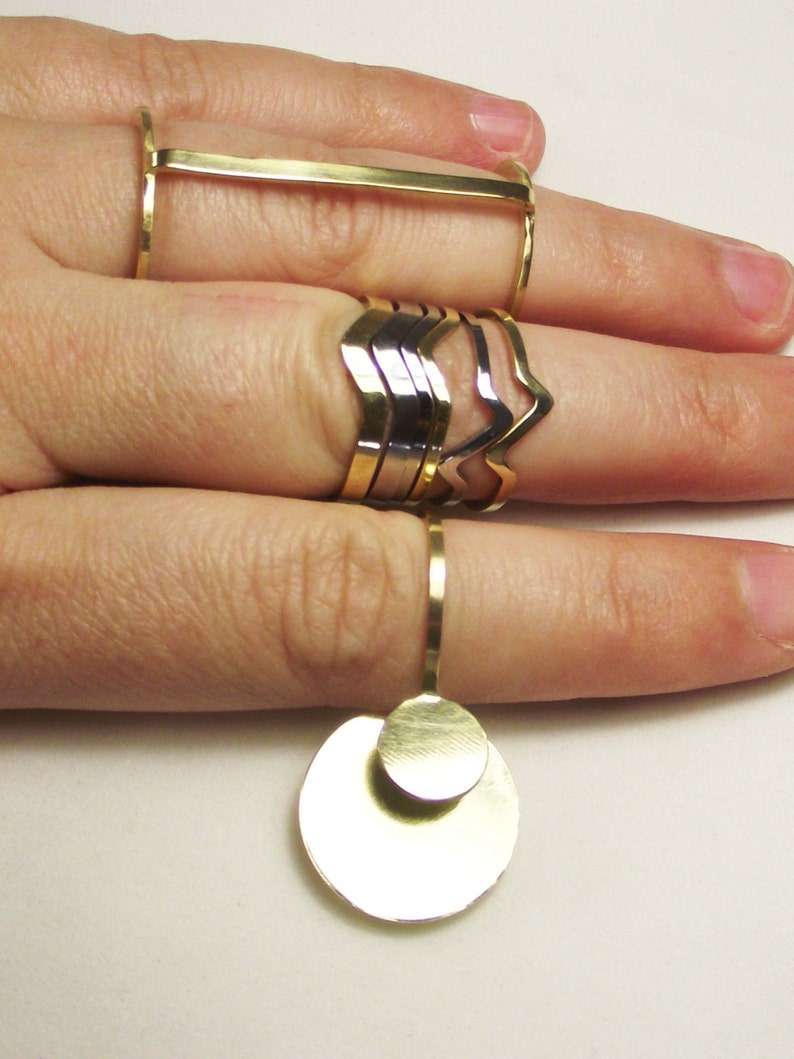May include: A set of gold and silver rings, including a ring with a large, round, double-layered pendant. The rings are stacked on a finger.
