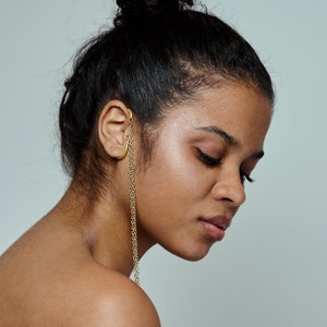 Può includere: Una donna con i capelli scuri raccolti in uno chignon indossa un orecchino a catena dorato.