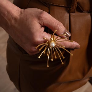 以下が含まれることがあります： 真珠が中央にあり、放射状の線が花火のようにデザインされた金の指輪です。