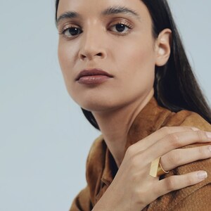 May include: A gold-toned ring featuring a wide, flat, rectangular front plate with a brushed metal finish. The band is smooth and circular, designed to sit comfortably on the finger. The piece has a clean, geometric aesthetic with sharp edges.