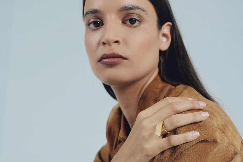 May include: A gold-toned ring featuring a wide, flat, rectangular front plate with a brushed metal finish. The band is smooth and circular, designed to sit comfortably on the finger. The piece has a clean, geometric aesthetic with sharp edges.