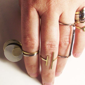 May include: A hand wearing a variety of gold and silver rings. The rings are stacked on the index finger and include a geometric design, a simple band, and a ring with a vertical bar.