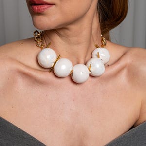 May include: A statement necklace featuring five large, smooth, off-white spherical beads with a subtle marbled finish. The beads are separated by thin, vertical gold-toned metal bars and connected to a thick gold-toned chain link necklace.